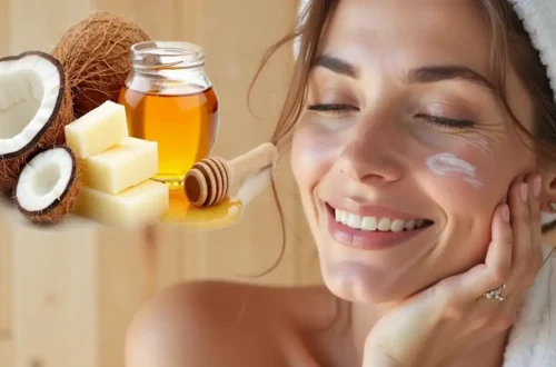 uma mulher jovem de pele clara e aparência saudável, com os olhos fechados e um sorriso relaxado no rosto. Ela usa uma toalha enrolada na cabeça, como se tivesse acabado de sair do banho, e apoia o rosto suavemente na mão direita. Sobre sua bochecha esquerda há uma pequena quantidade de creme branco, aplicada em forma de espiral. No lado esquerdo da imagem, há uma montagem de ingredientes naturais em destaque: um coco partido ao meio mostrando sua polpa branca, pedaços de manteiga vegetal (possivelmente manteiga de karité), um pote de vidro com mel dourado e uma colher de pau usada para mel. O fundo é de madeira clara, transmitindo uma sensação de aconchego e naturalidade.